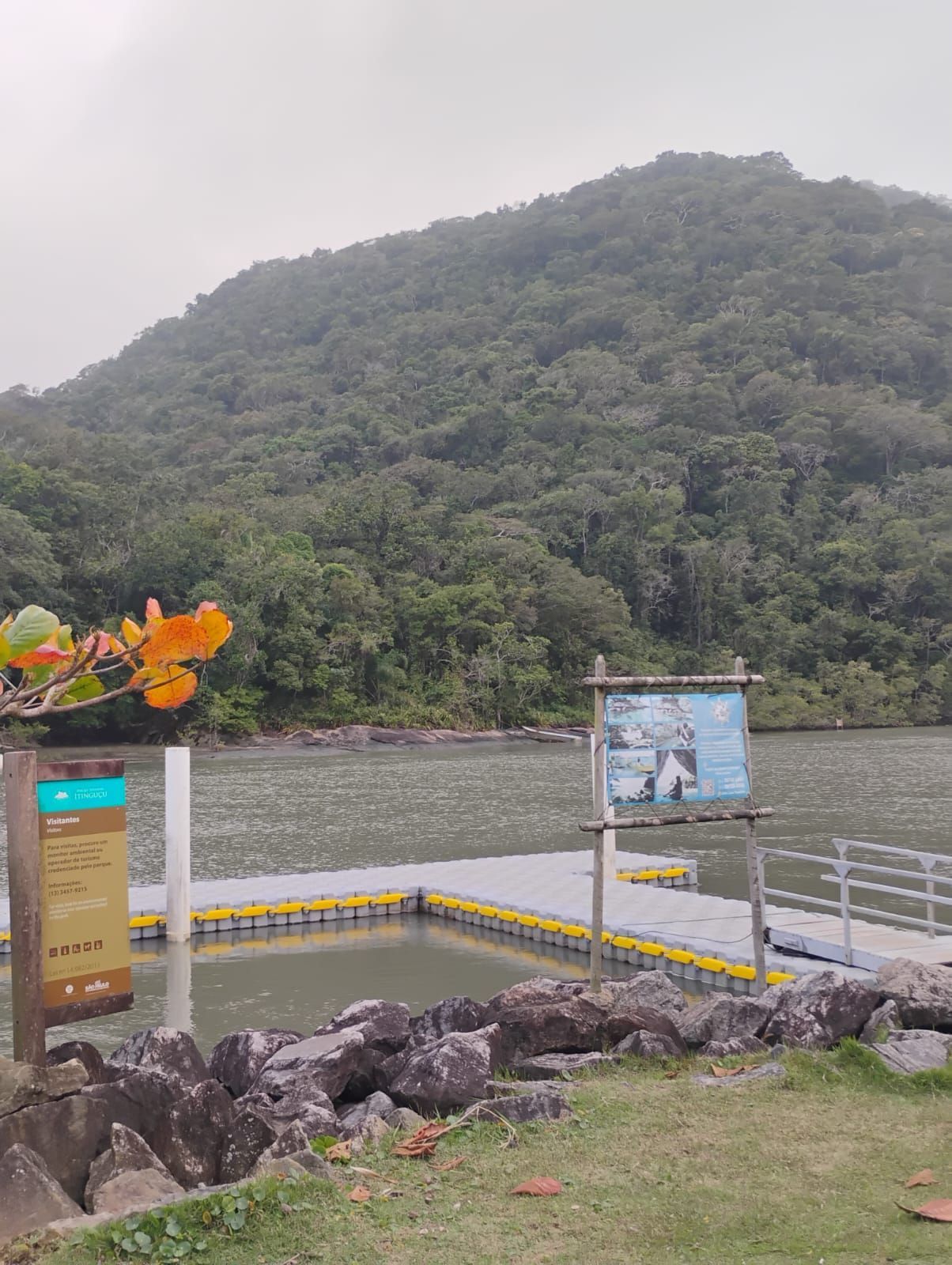 Um lago com um cais flutuante, placas e uma montanha ao fundo sob um céu nublado.