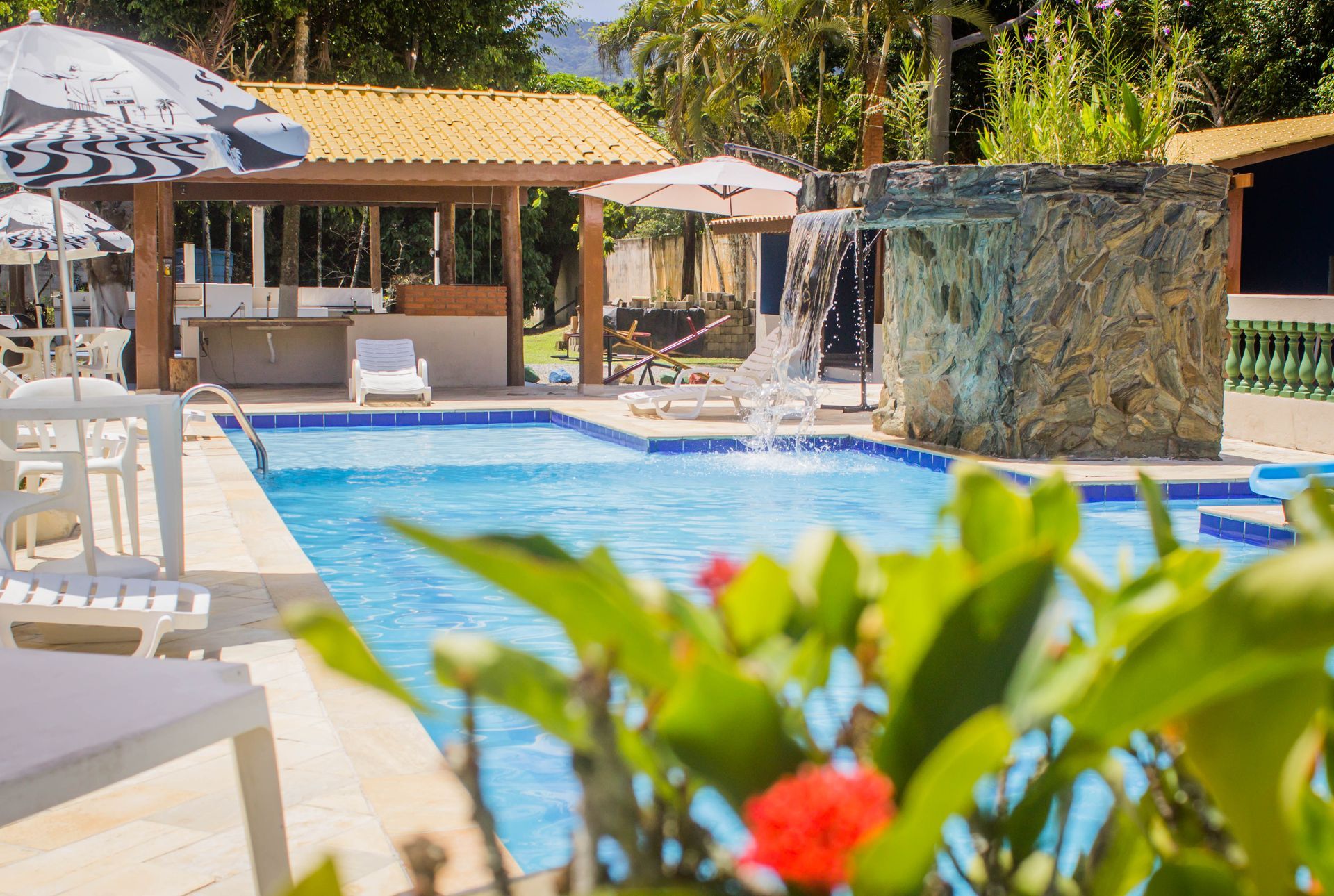 Piscina com uma pequena cascata, espreguiçadeiras e cozinha ao ar livre.