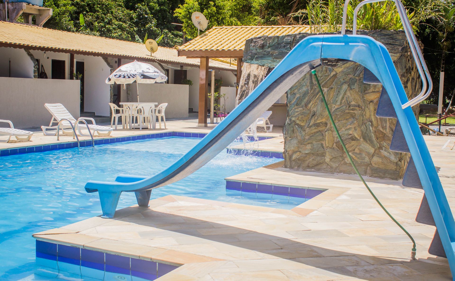 Piscina com toboágua azul, espreguiçadeiras e prédios ao fundo. Ambiente externo ensolarado.