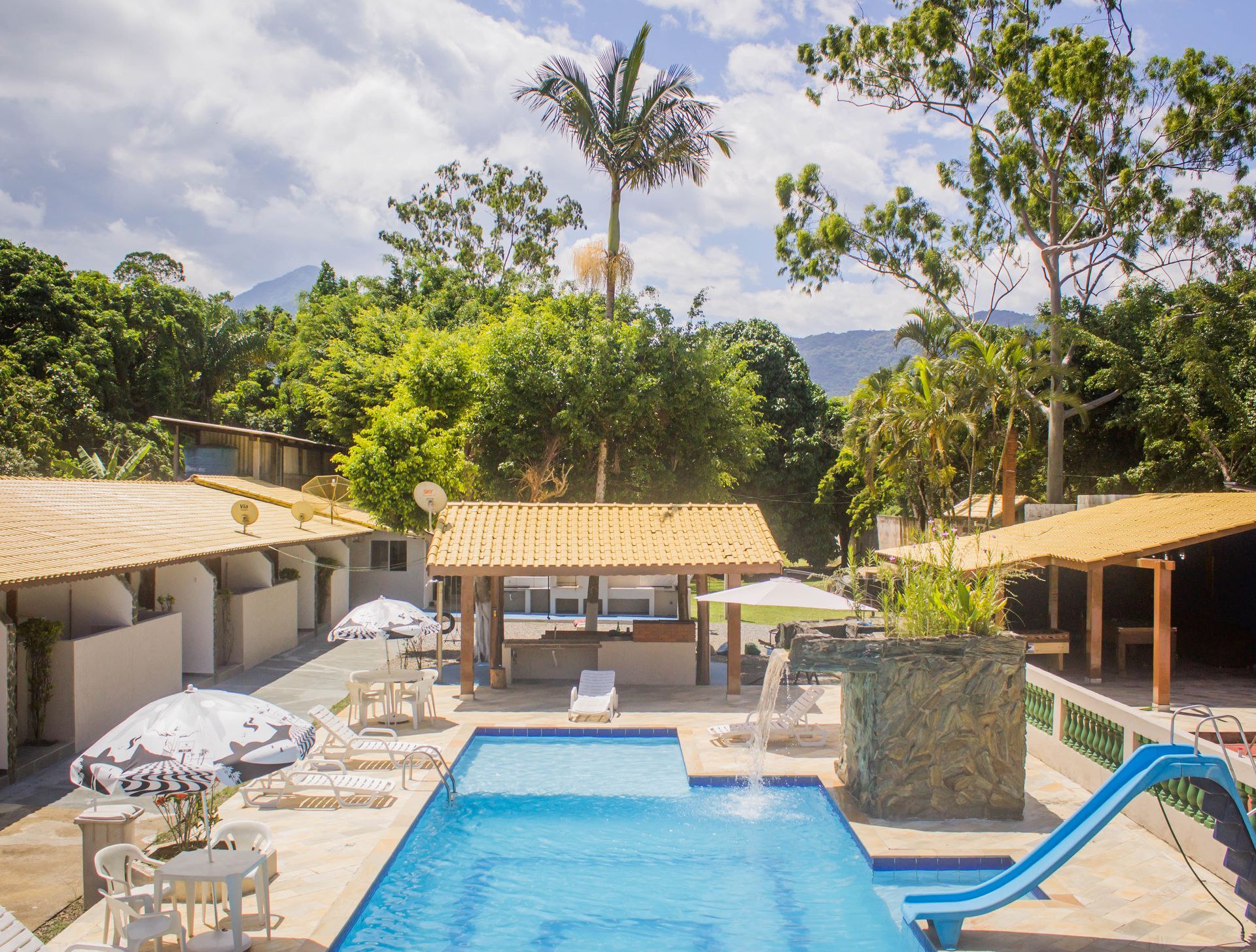 Piscina em um resort com bangalôs, guarda-sóis, um escorregador e uma área de bar cercada por árvores verdejantes.