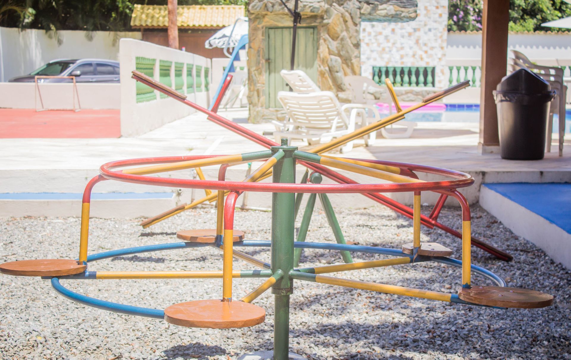 Carrossel com postes vermelhos, amarelos e azuis, assentos em um playground e uma piscina ao fundo.