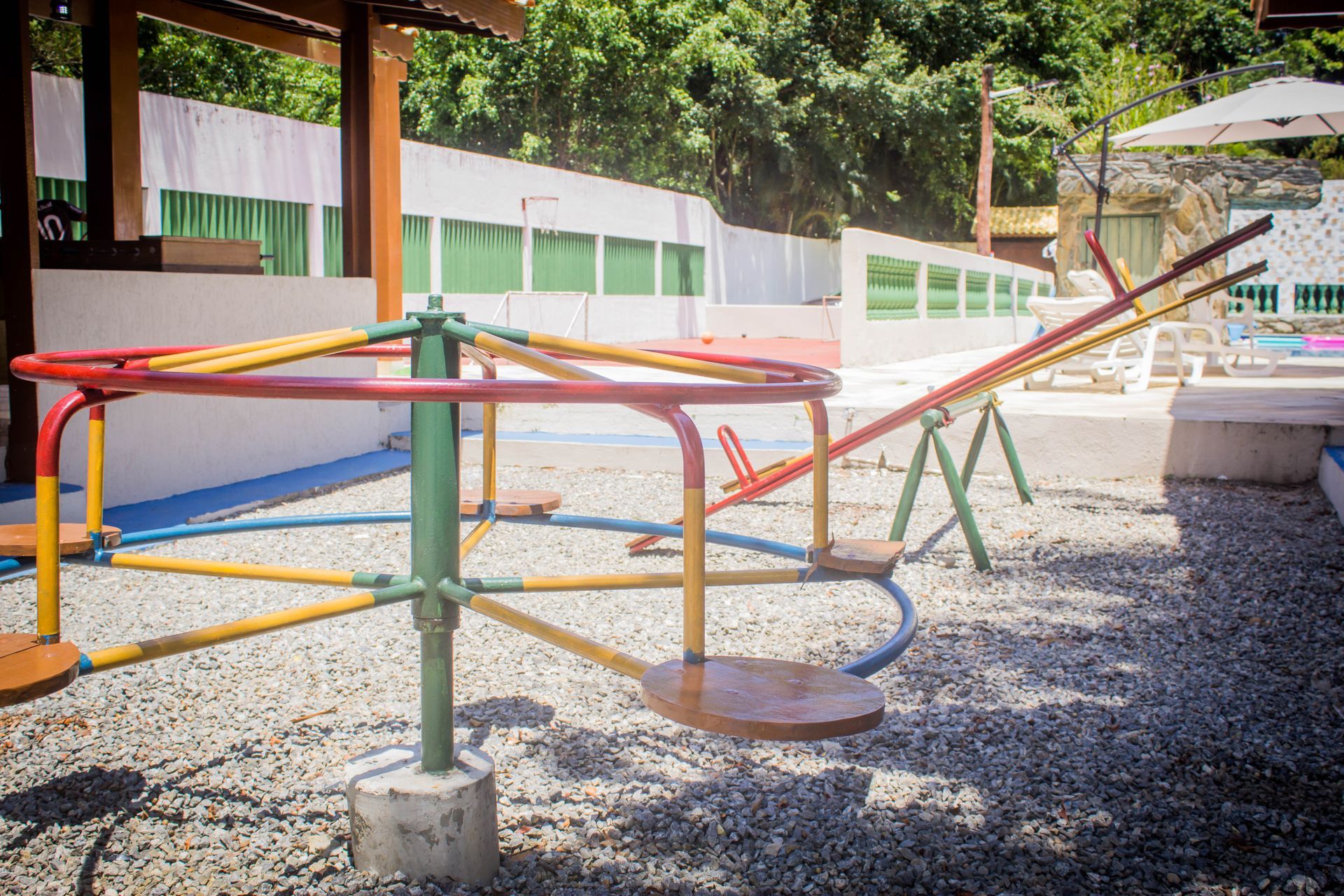 Parque infantil com carrossel colorido e gangorra em terreno de cascalho perto de cerca branca e piscina.
