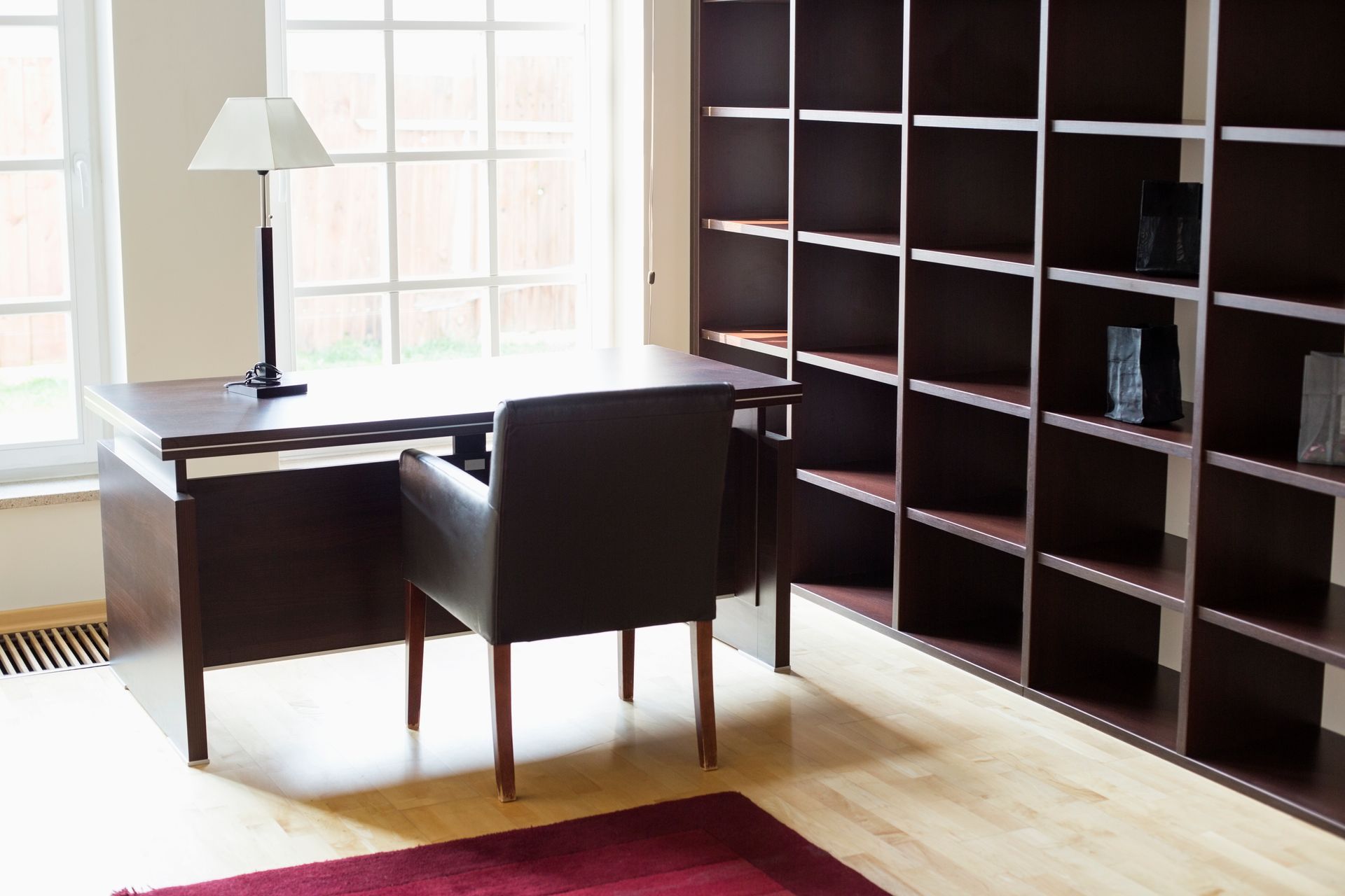 A home office with a dark desk, chair, and bookshelf. A lamp sits on the desk near a window.