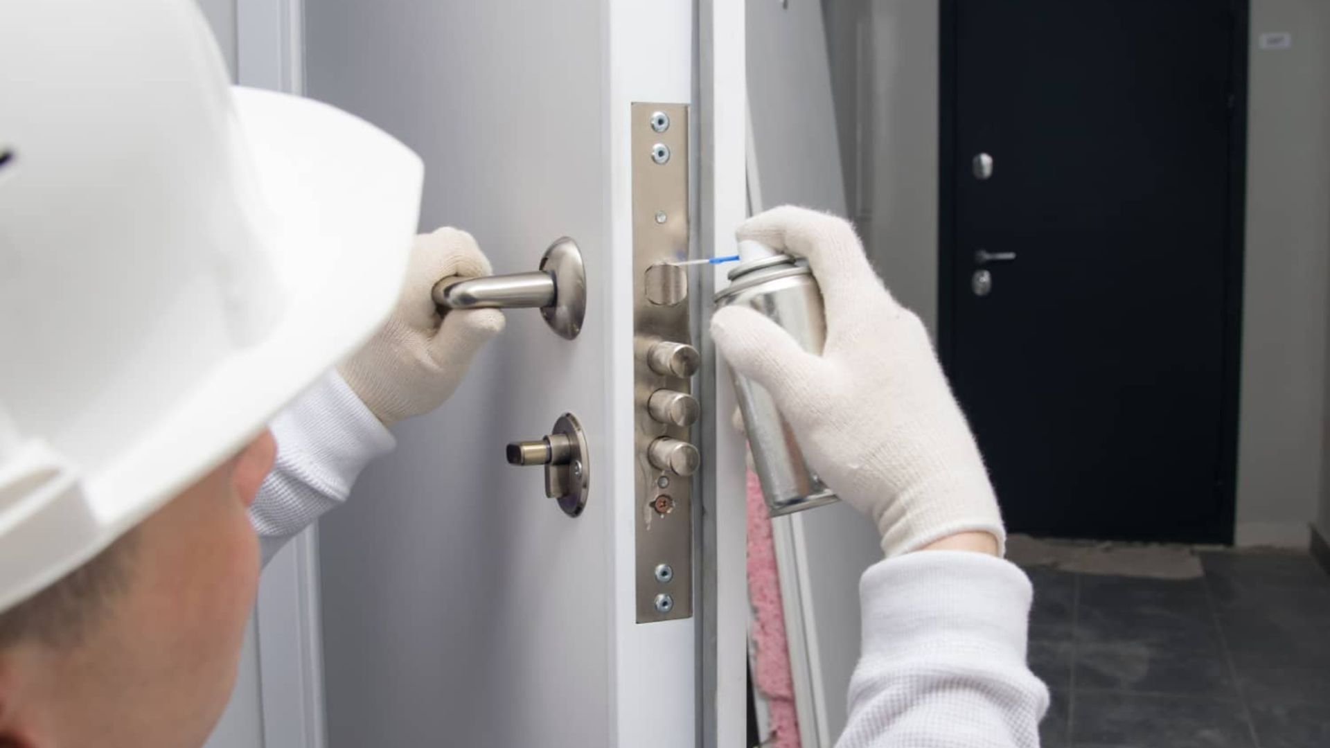 Gloved hands installing a silver door lock on a white door with a drill in a hallway