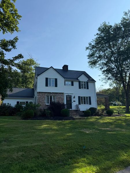 White two-story house with black roof and brick lower level, surrounded by a grassy yard and trees under a blue sky