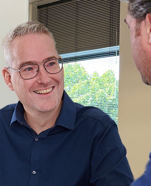 Een man met een bril lacht terwijl hij met een andere man praat.