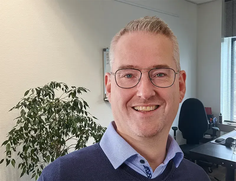 Een glimlachende Coen in een blauw overhemd staat in een kantoor, met een plant en een bureau op de achtergrond.