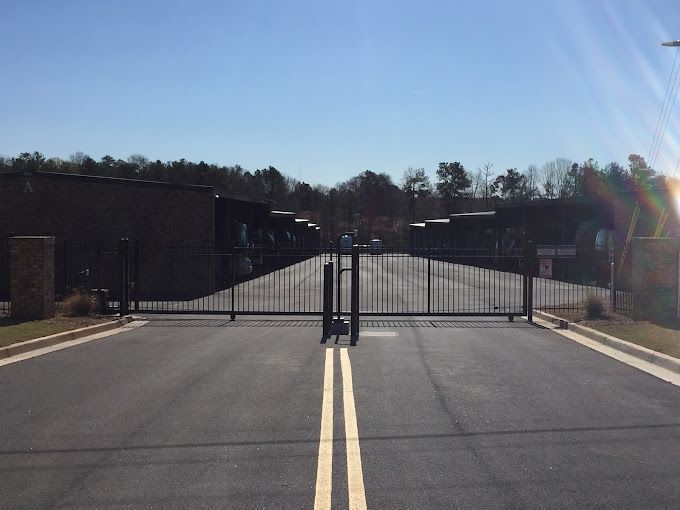 Black security gate across a paved road, leading into a facility with storage units; sunny day.
