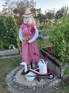 A scarecrow in a pink dress is standing in a garden next to a wheelbarrow.