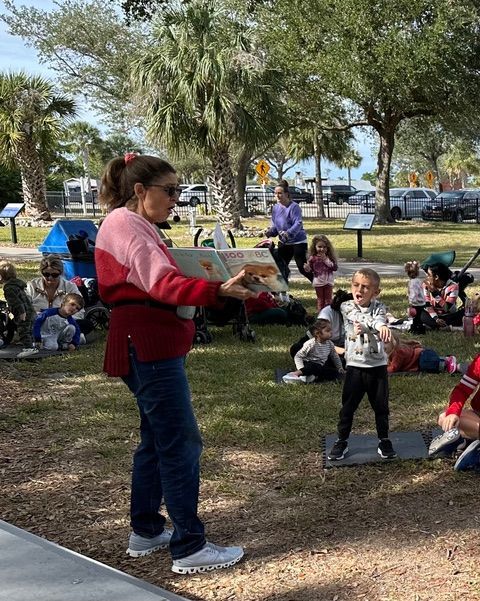 A woman is reading a book to a group of children in a park.