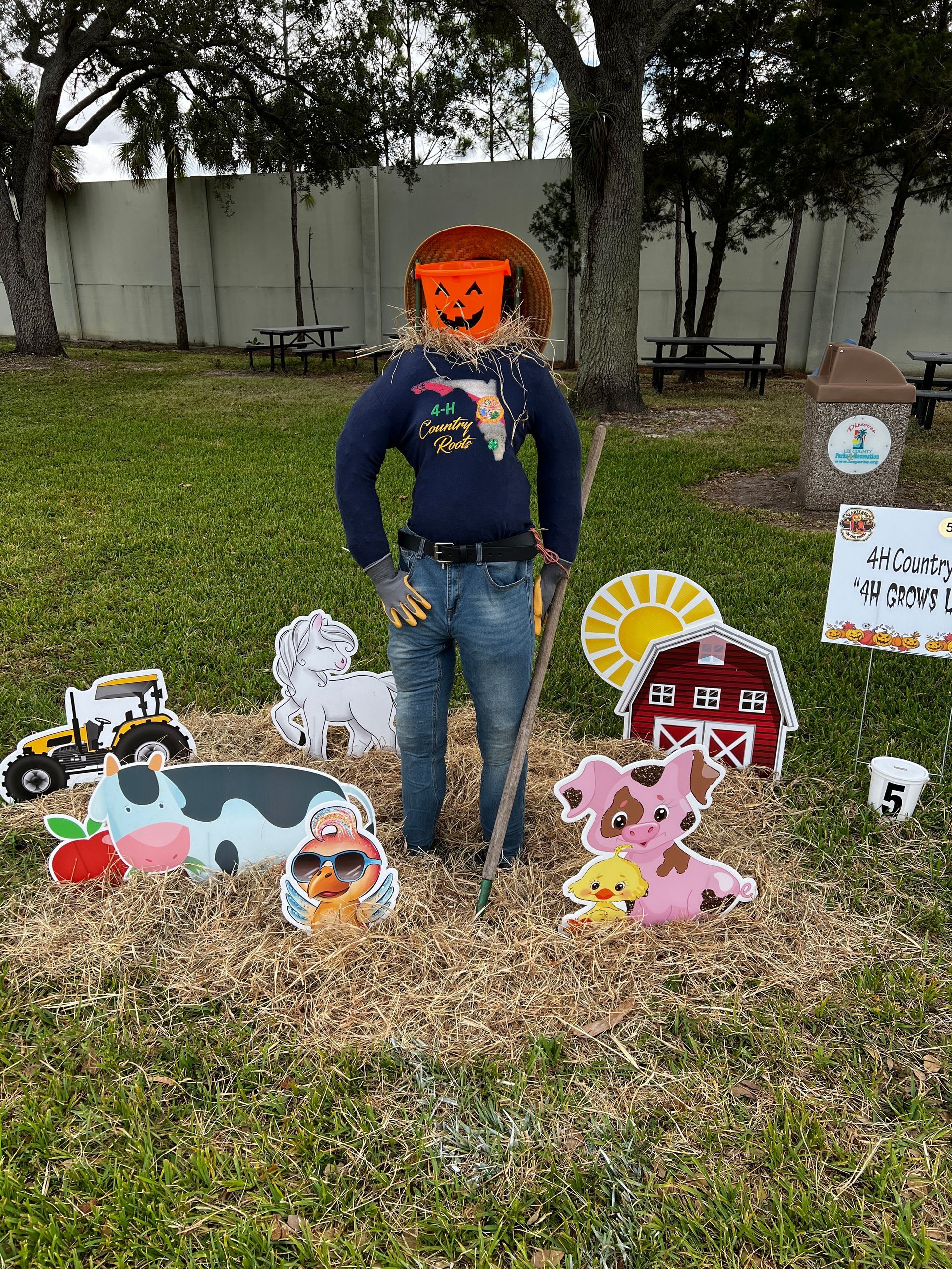 A scarecrow with a pumpkin on his face is standing in the grass surrounded by farm animals.
