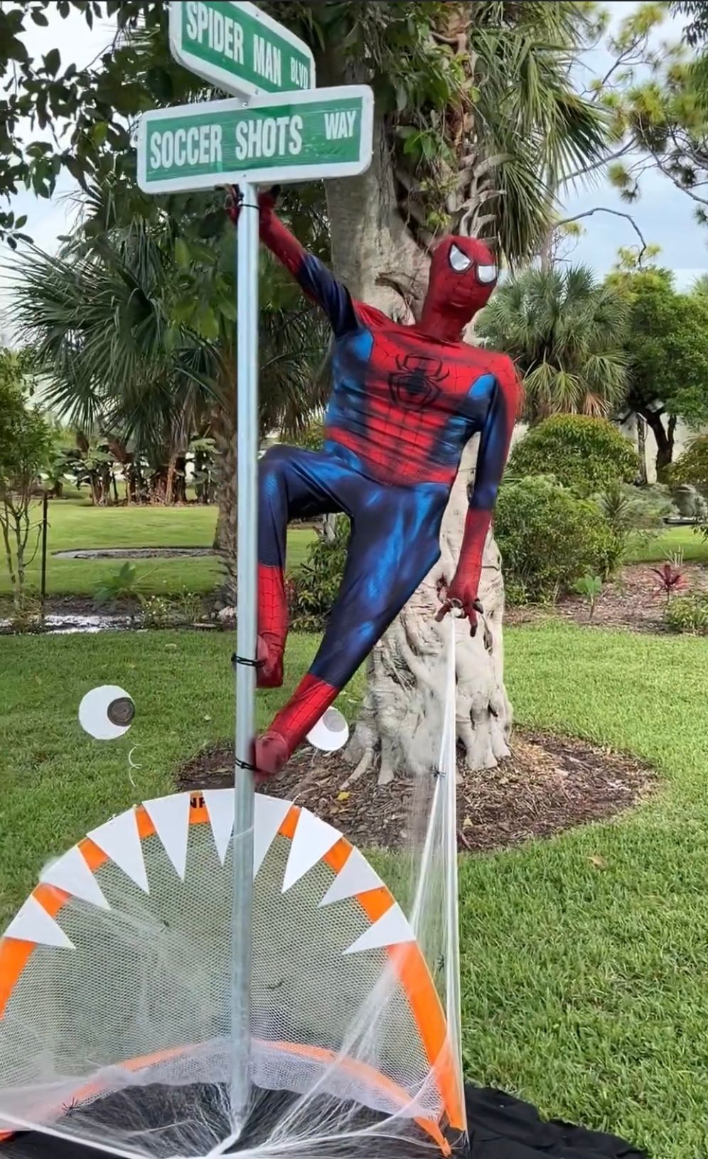 A spiderman statue is hanging from a street sign in a park.