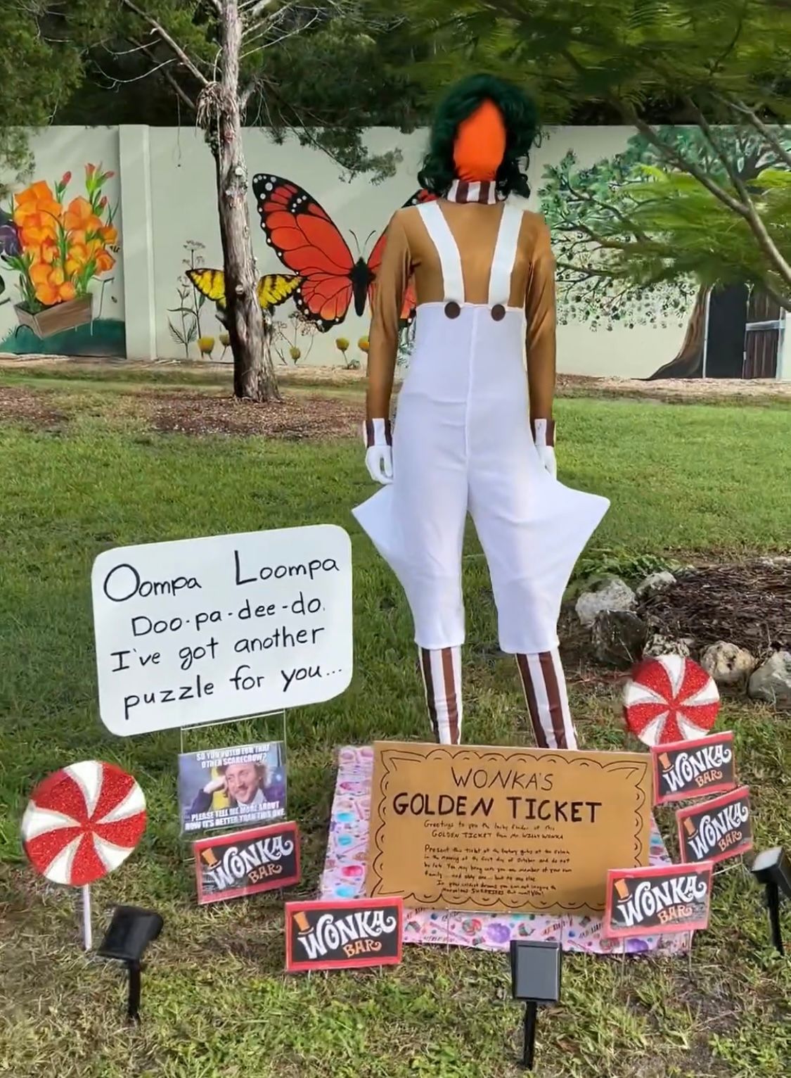 A statue of willy wonka is standing in the grass next to a sign.