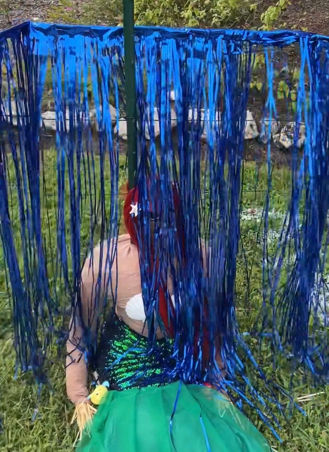 A woman in a green dress is sitting in the grass behind a blue curtain.
