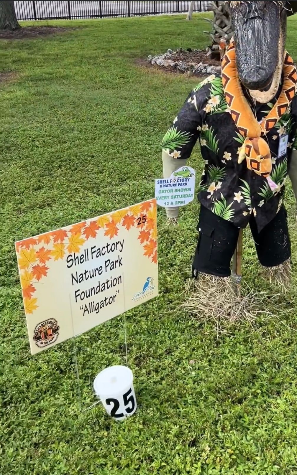 A scarecrow in a hawaiian shirt is standing in the grass next to a sign.