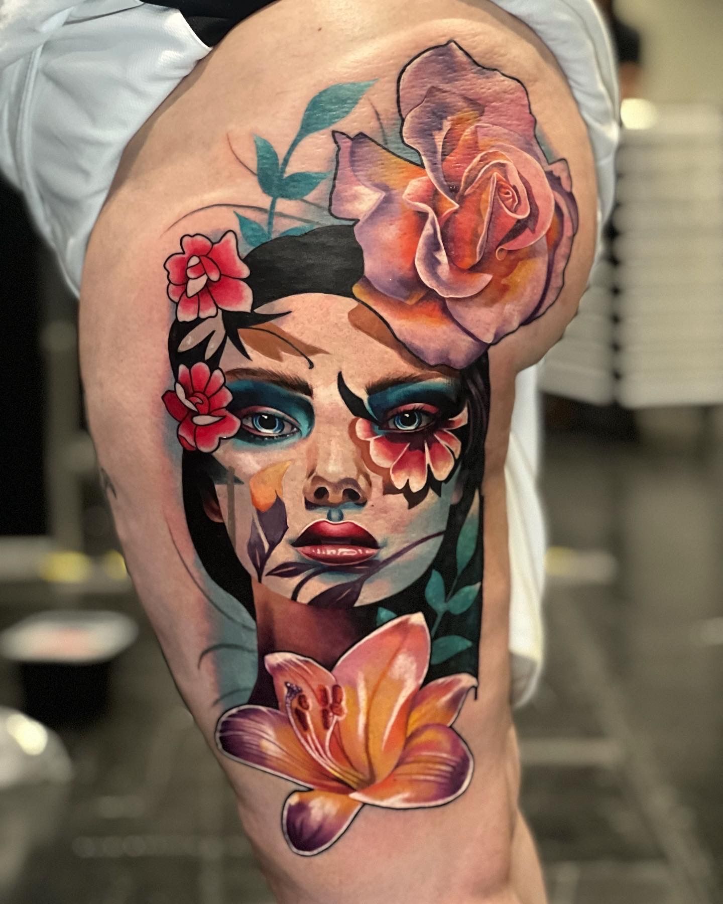 Tattoo of a woman's face with colorful floral designs on a thigh. The portrait has intense blue eyes, and a large rose is behind her head.