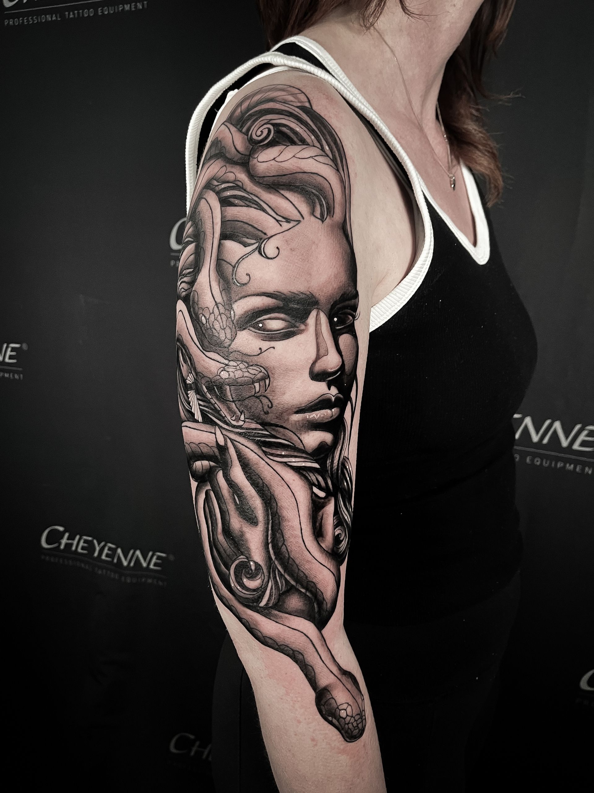 Woman's arm tattooed with a detailed black and white portrait, possibly a mythological figure. She wears a black tank top.