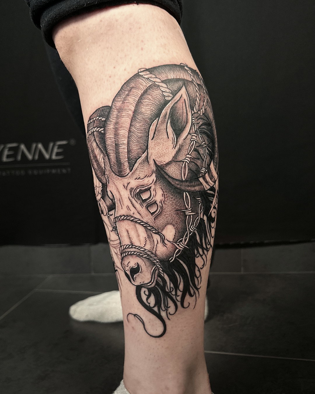 Black and grey calf tattoo of a ram's head with horns, wearing a mask and chains.