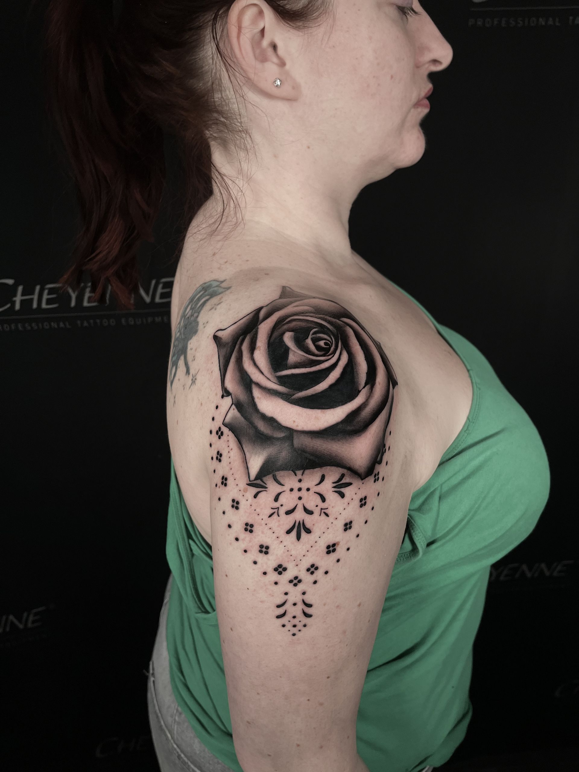 Woman with a black and gray rose tattoo on her shoulder, wearing a green tank top. A pattern of dots extends from the rose.