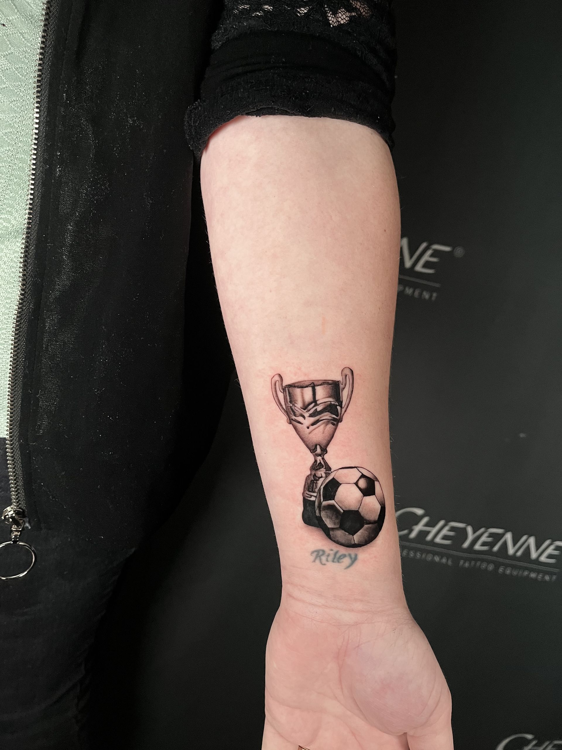 A black and grey tattoo on a forearm of a trophy and soccer ball. A name is below the ball.