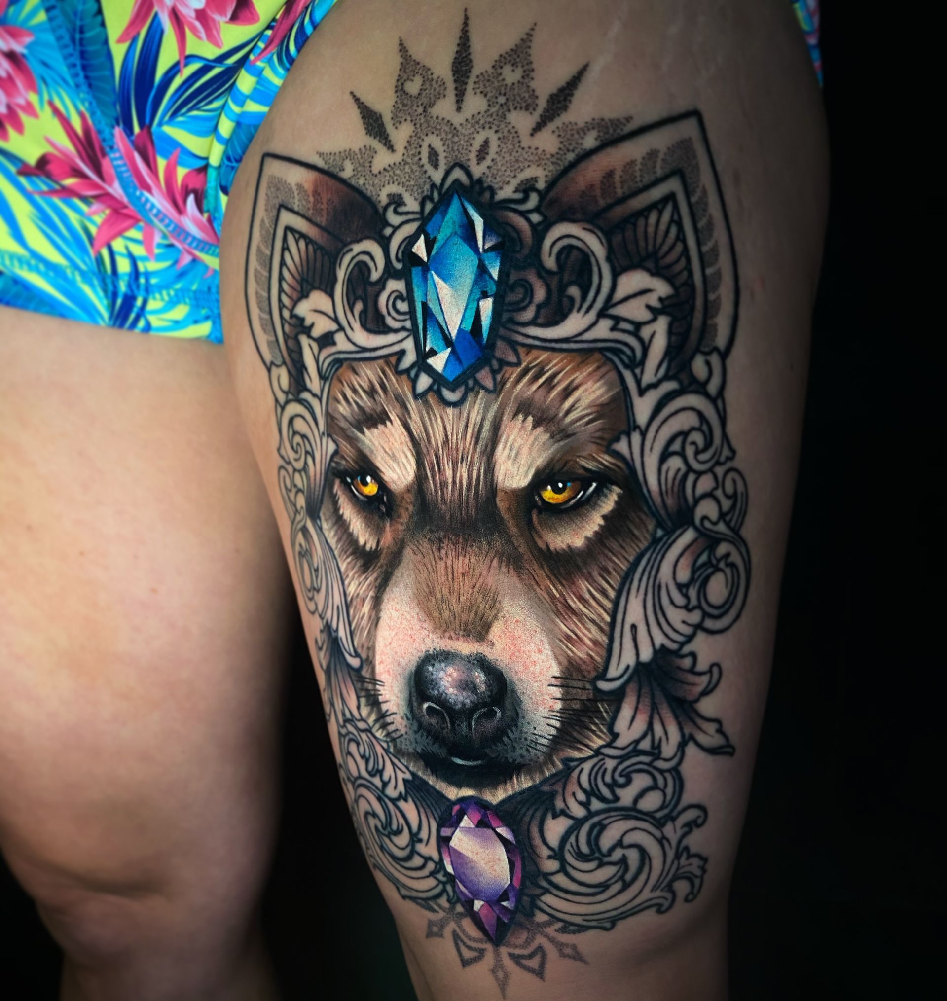 Tattoo of a wolf's head with a jewel-encrusted crown, set against ornate swirls, on a person's thigh. The wolf has yellow eyes.
