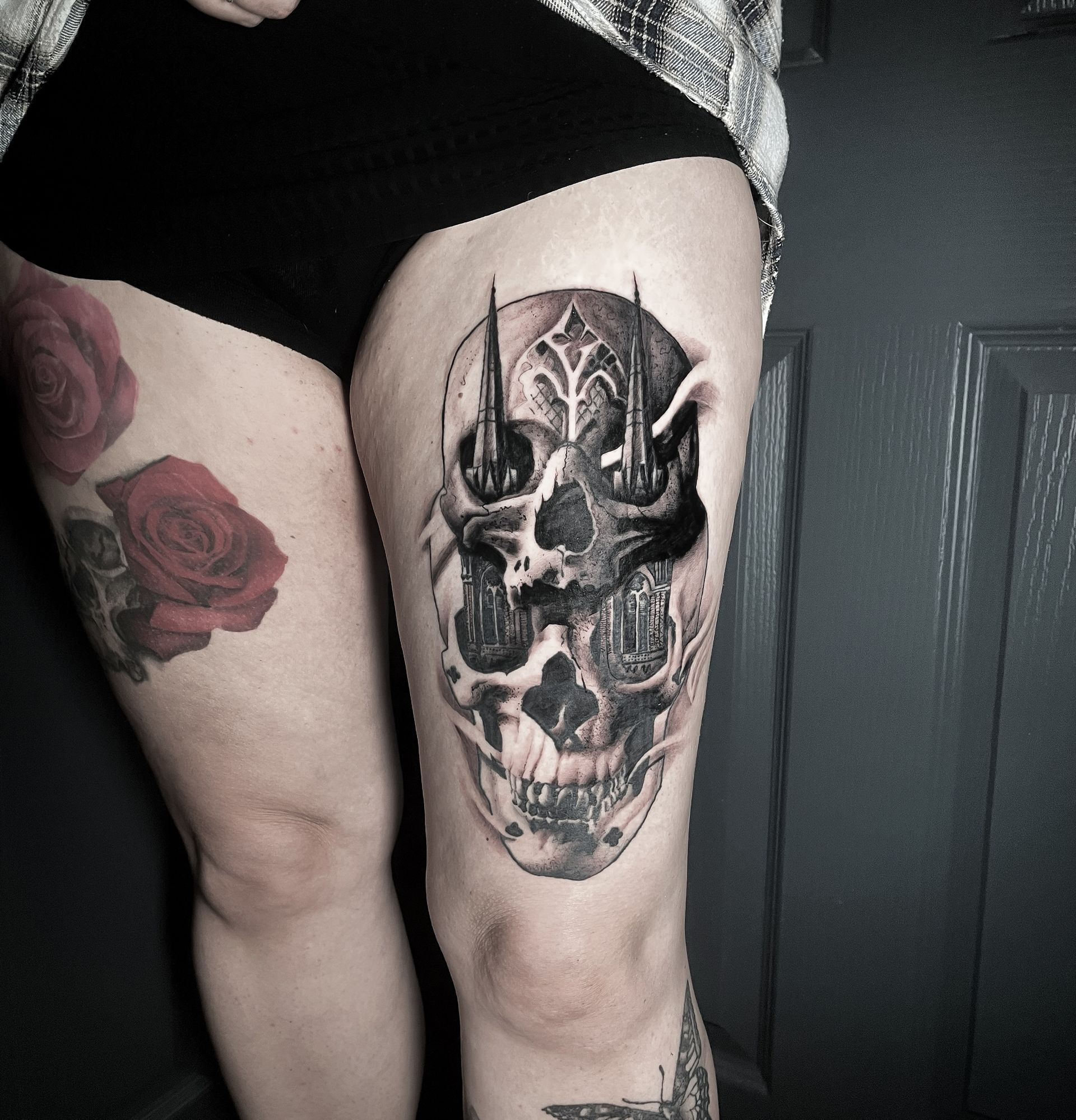 Woman's thigh with a detailed black and gray skull tattoo featuring gothic architectural elements; a red rose tattoo is visible on the other thigh.