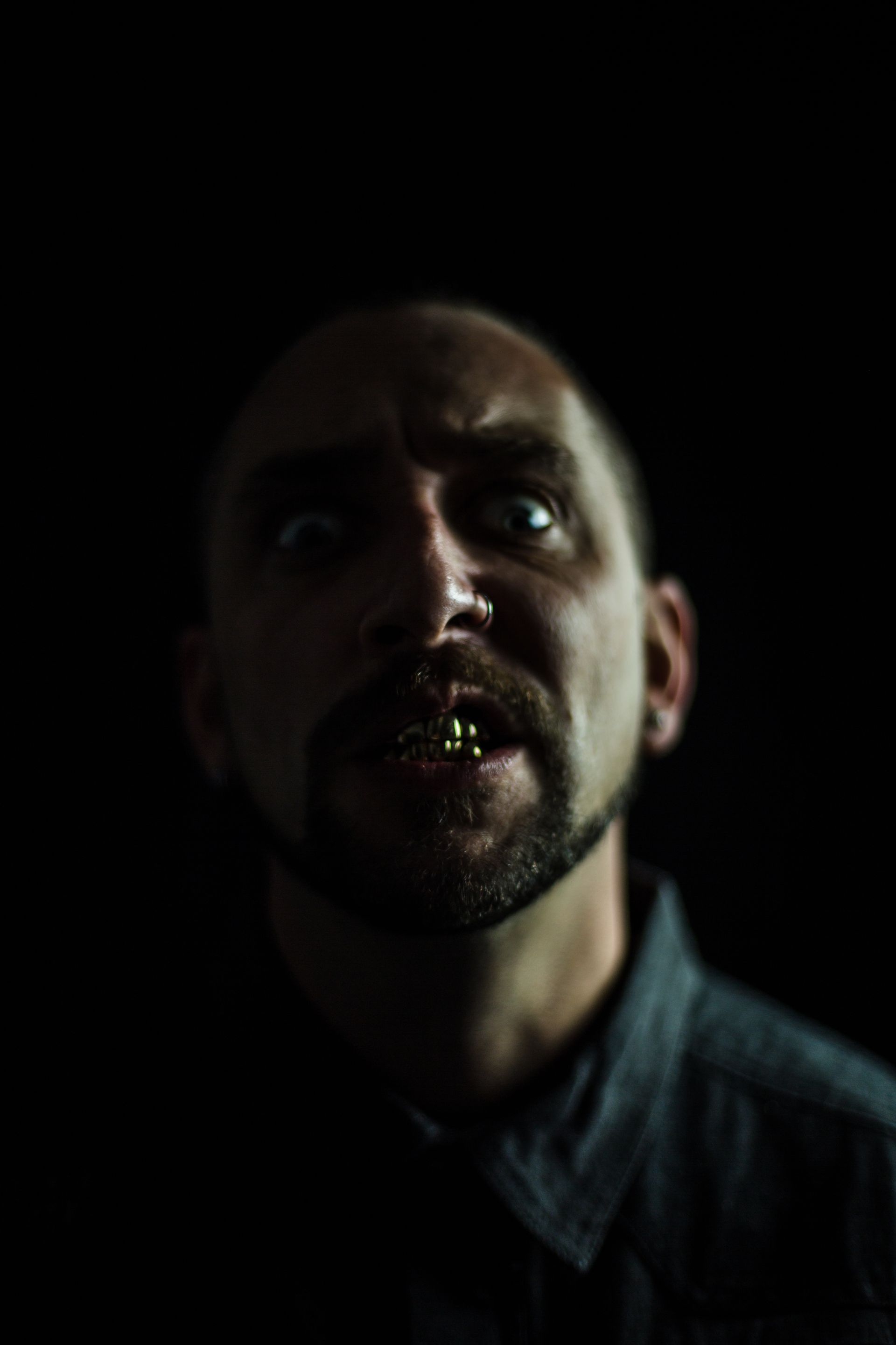 A man with a dark background, looking up with a sinister expression, mouth slightly open, showing teeth.