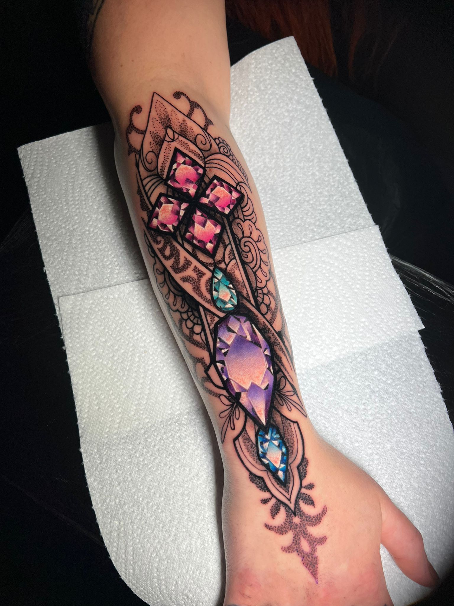 Arm tattoo featuring jewel-like designs in pink, purple, and teal, with intricate black filigree patterns.