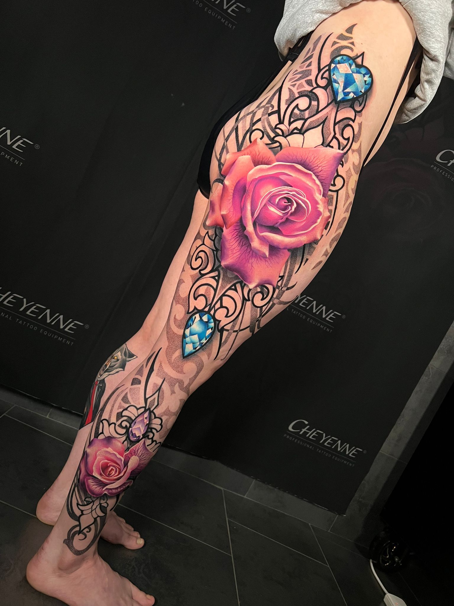 Leg tattoo featuring a large pink rose, jewels, and intricate black filigree designs, covering most of the leg.