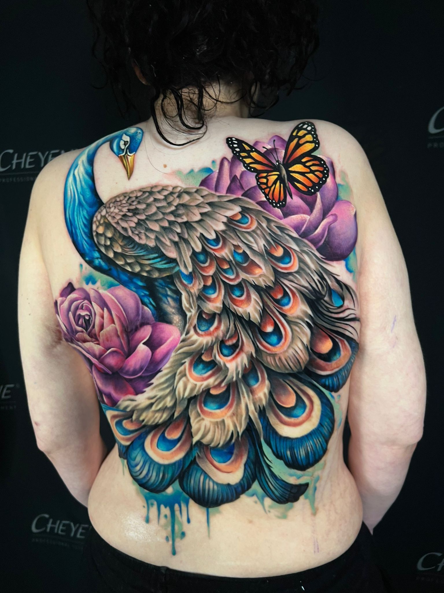 A vibrant peacock tattoo on a person's back, featuring blue, green, and orange feathers, surrounded by purple roses and a butterfly.