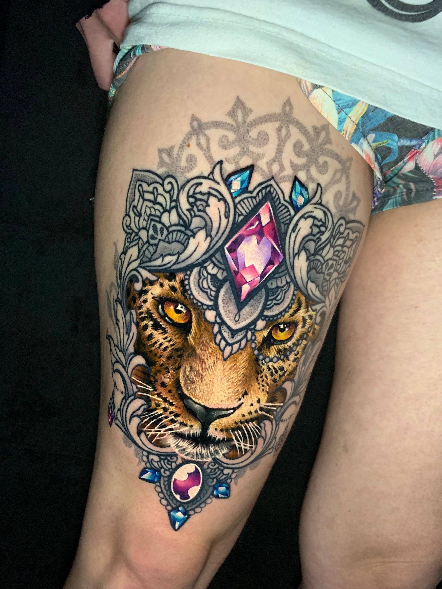A detailed tattoo of a leopard's face with jewel accents and ornate details on a person's thigh.