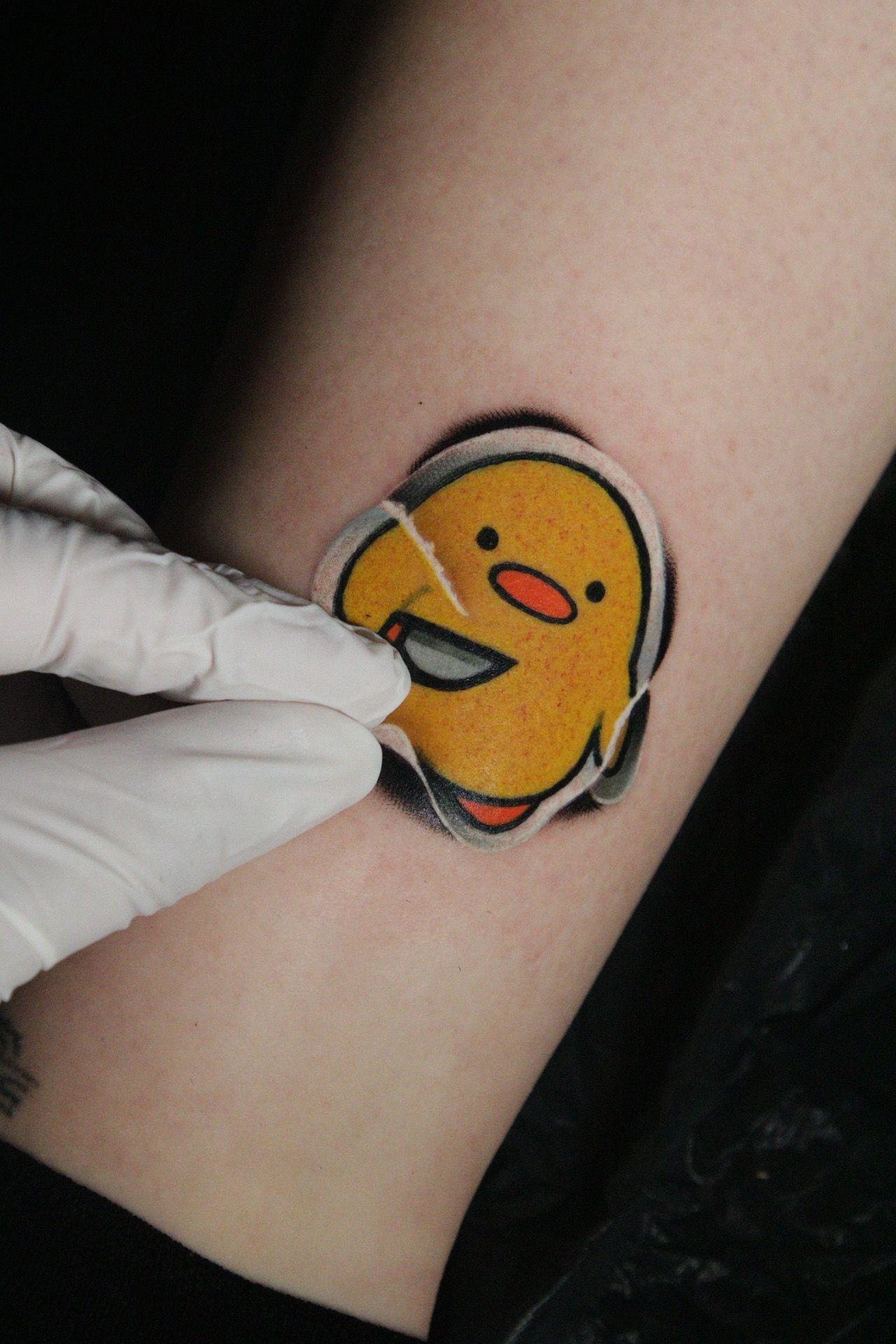 A person removes clear bandage from a fresh, colorful tattoo of a yellow cartoon chick with a knife. The tattoo is on an arm.