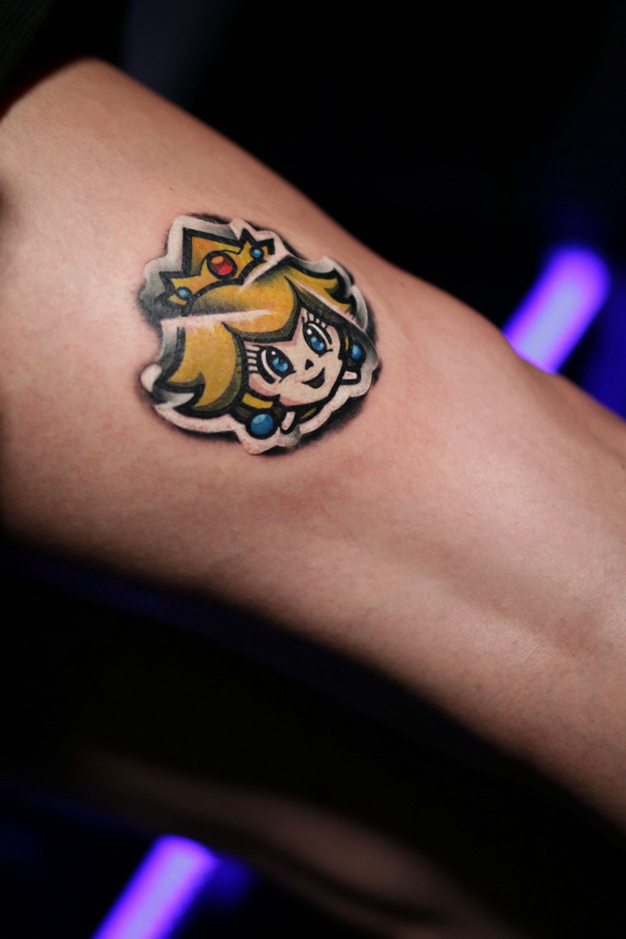 A colorful tattoo of Princess Peach from Super Mario on a person's arm, with her smiling face, crown, and blonde hair.