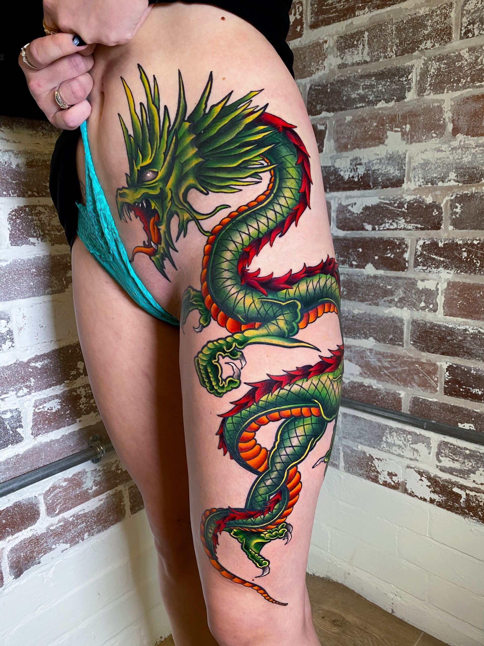 Dragon tattoo in green, red, and orange ink on a person's thigh and hip, posed against a brick wall.