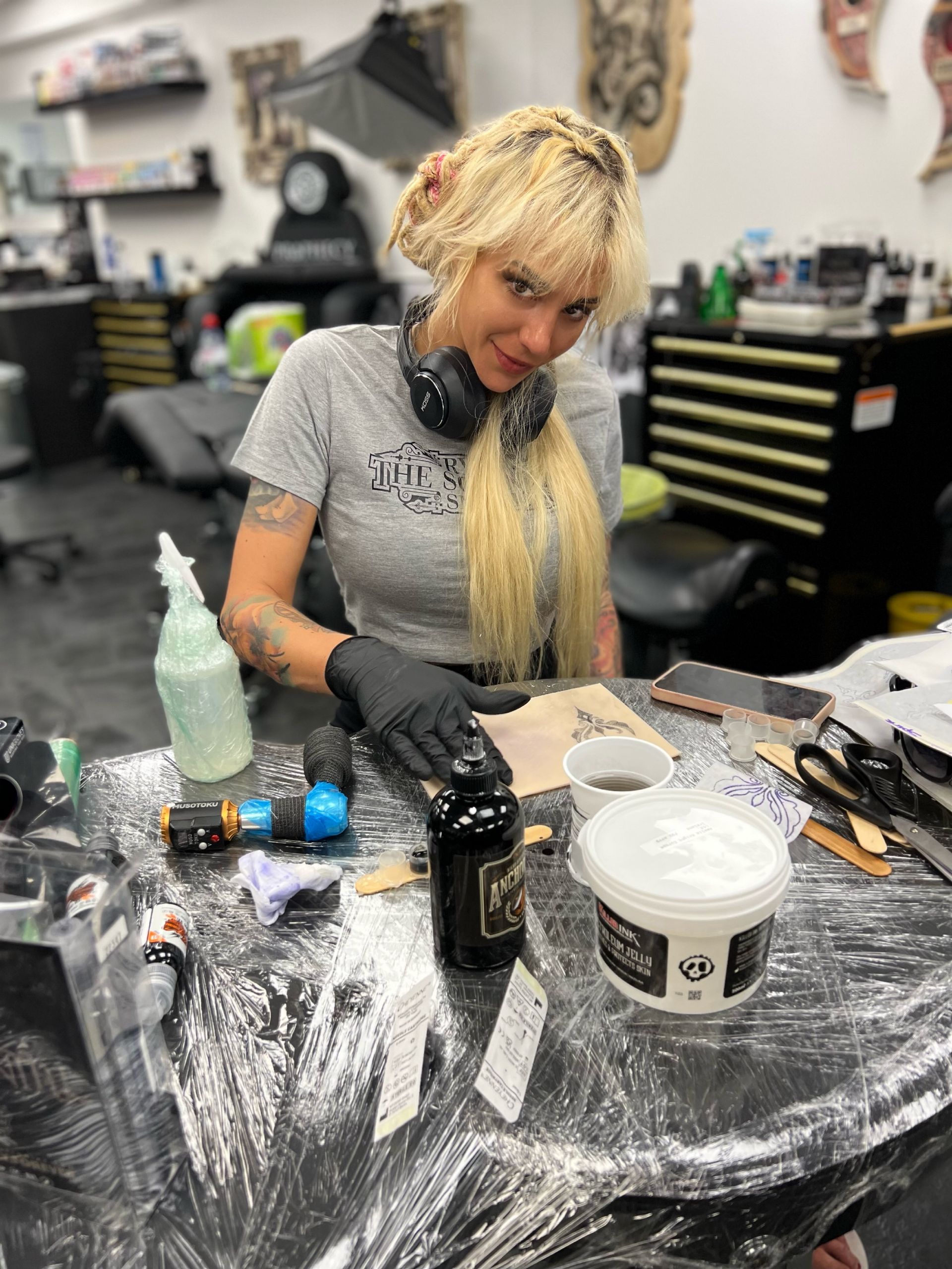Blonde woman in tattoo studio, preparing ink. She wears black gloves, a gray shirt, and smiles.