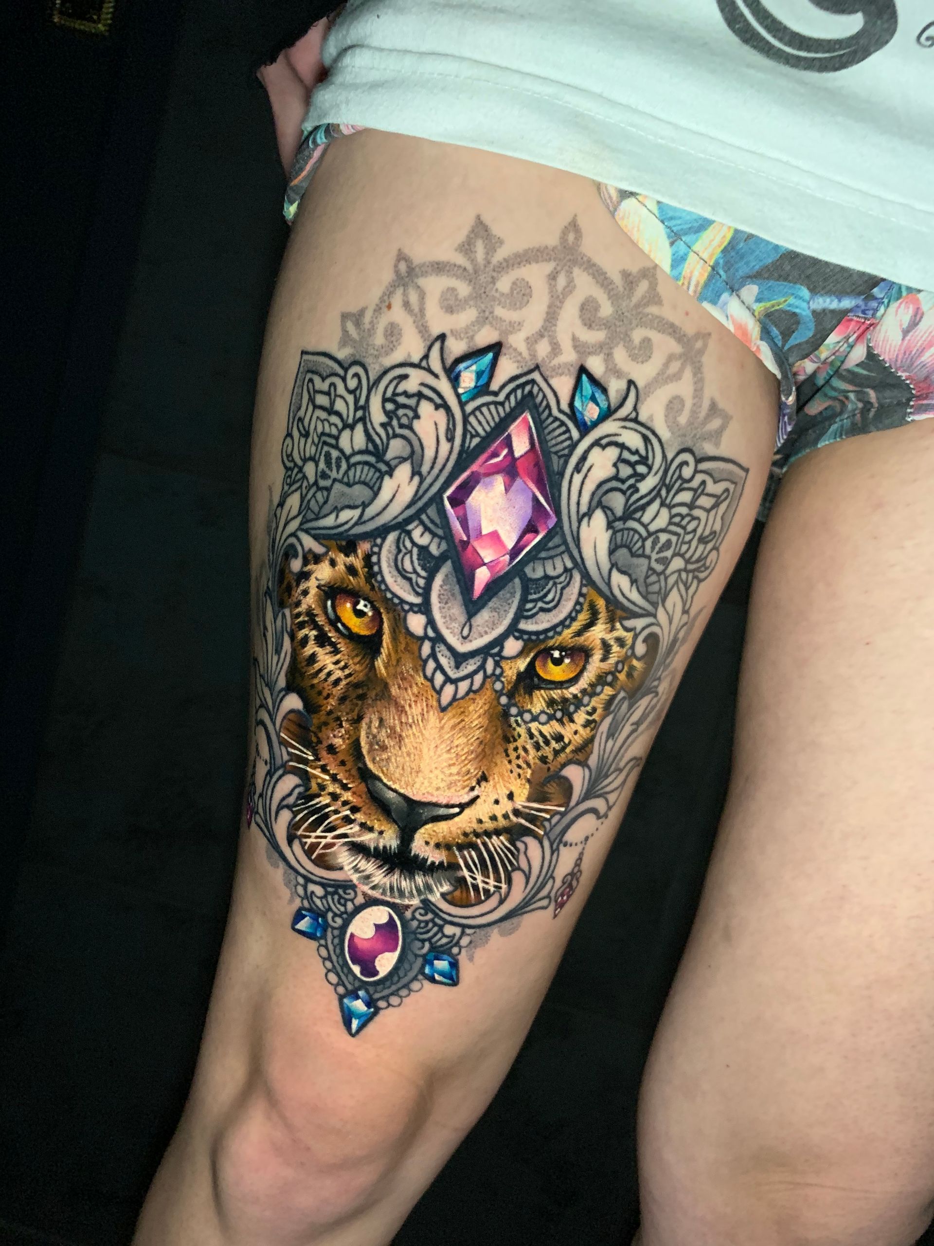 Tattoo of a leopard's face adorned with jewels and ornate scrollwork on a person's upper thigh.