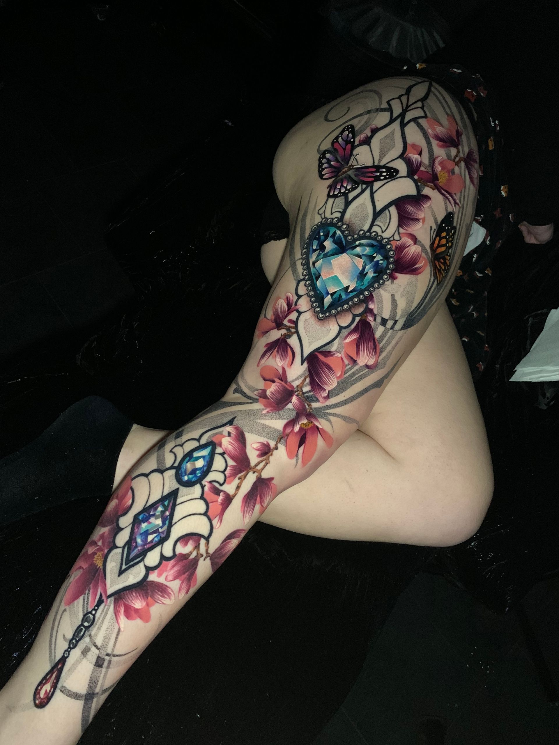 Tattoo of a jeweled heart and teardrop surrounded by pink flowers and butterflies on a person's leg.