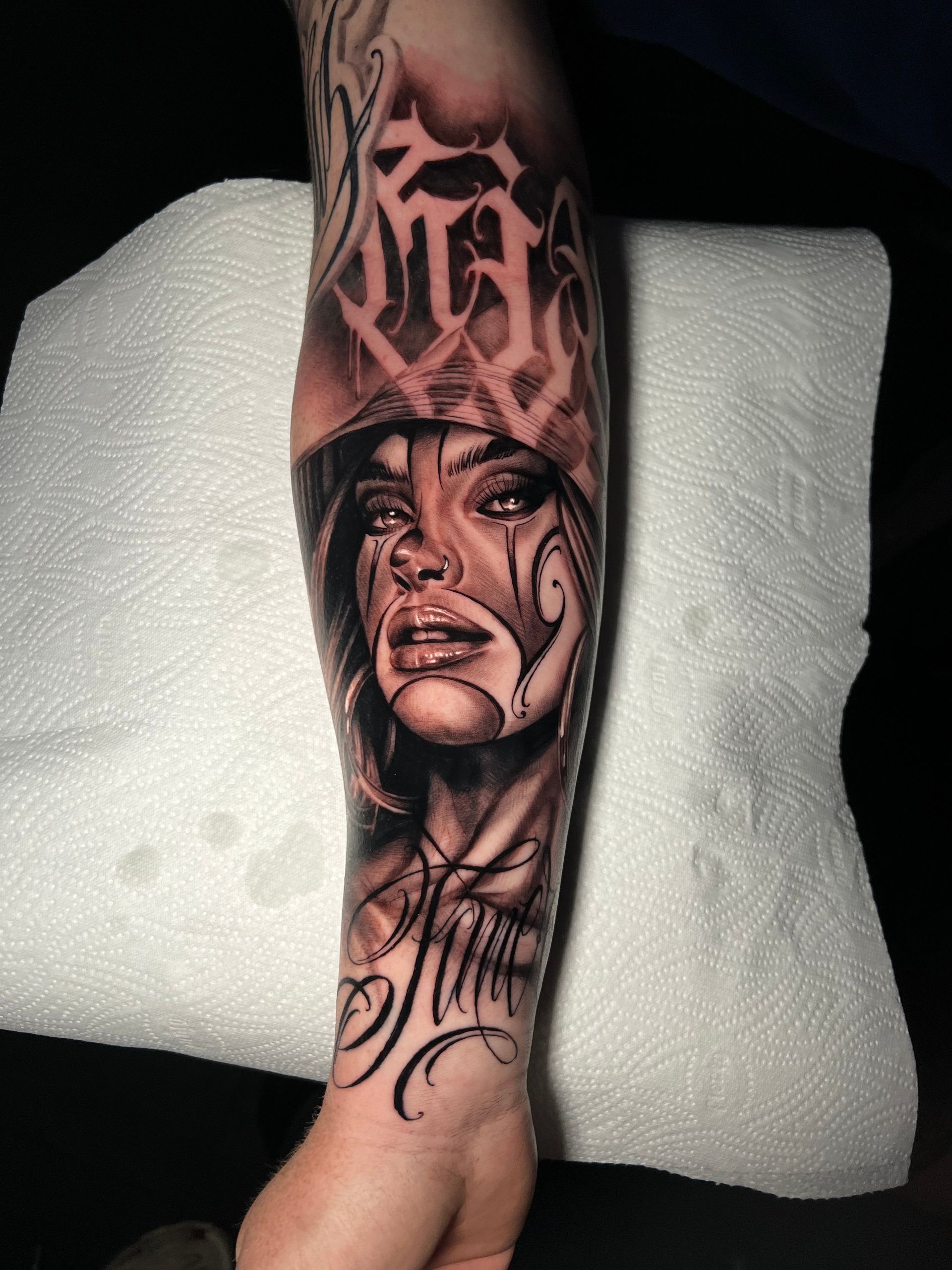 Black and grey tattoo on arm of a woman's face with clown-like makeup, a hat, and script above and below the face.