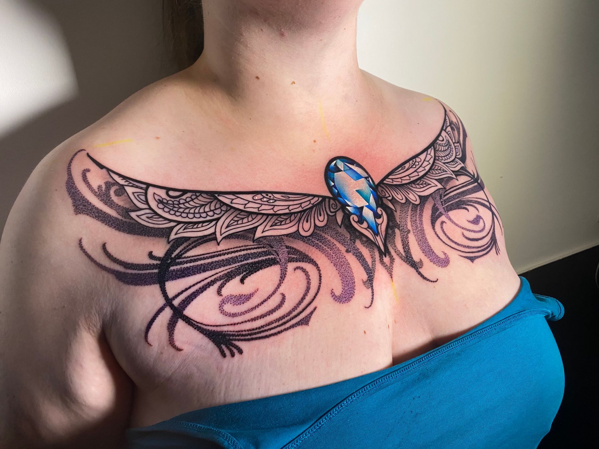 Tattoo on a person's chest and shoulder; features a blue gem with wing-like designs and swirling patterns, inked in black and gray.