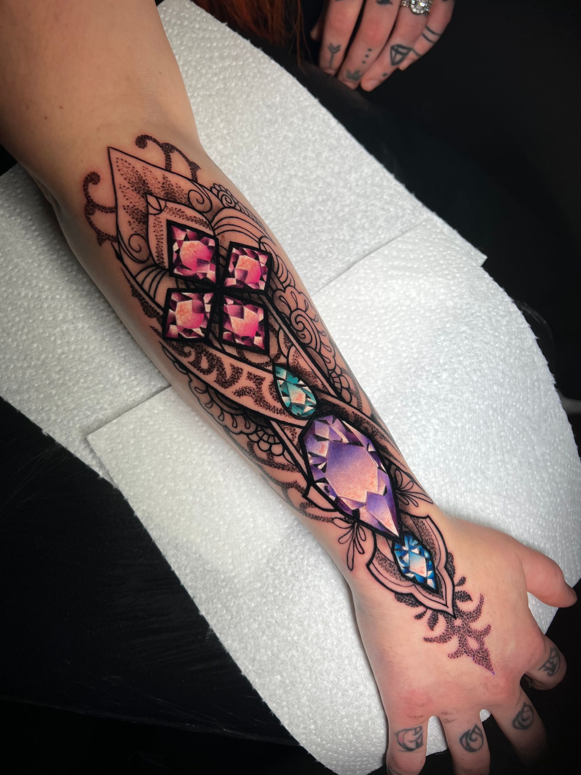 Tattoo of ornate jewel design on a person's arm. The jewels are pink, purple, and blue with intricate black line work.