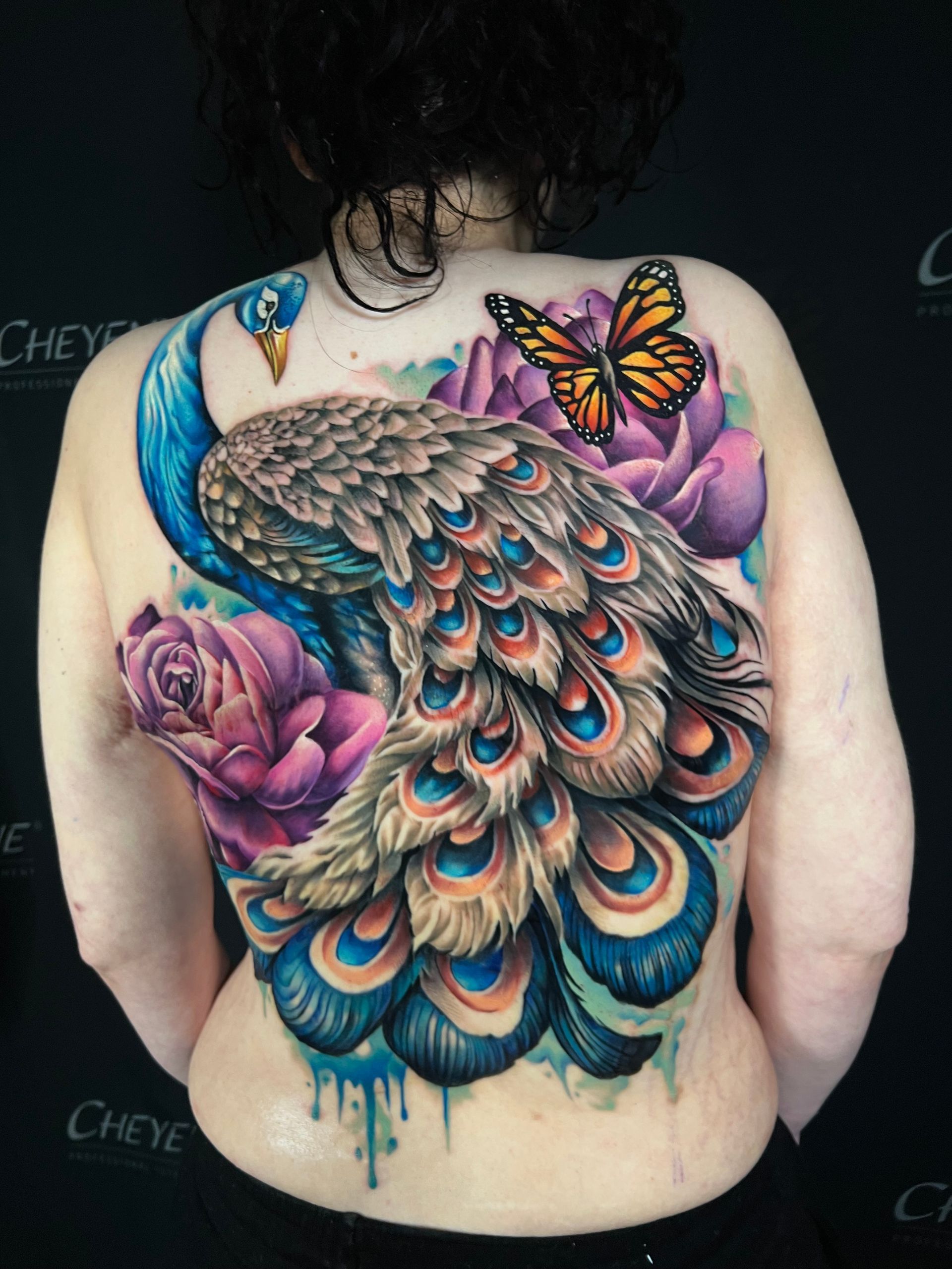 Tattoo of a vibrant peacock with a flowing tail, surrounded by pink and purple flowers and a butterfly on a person's back.