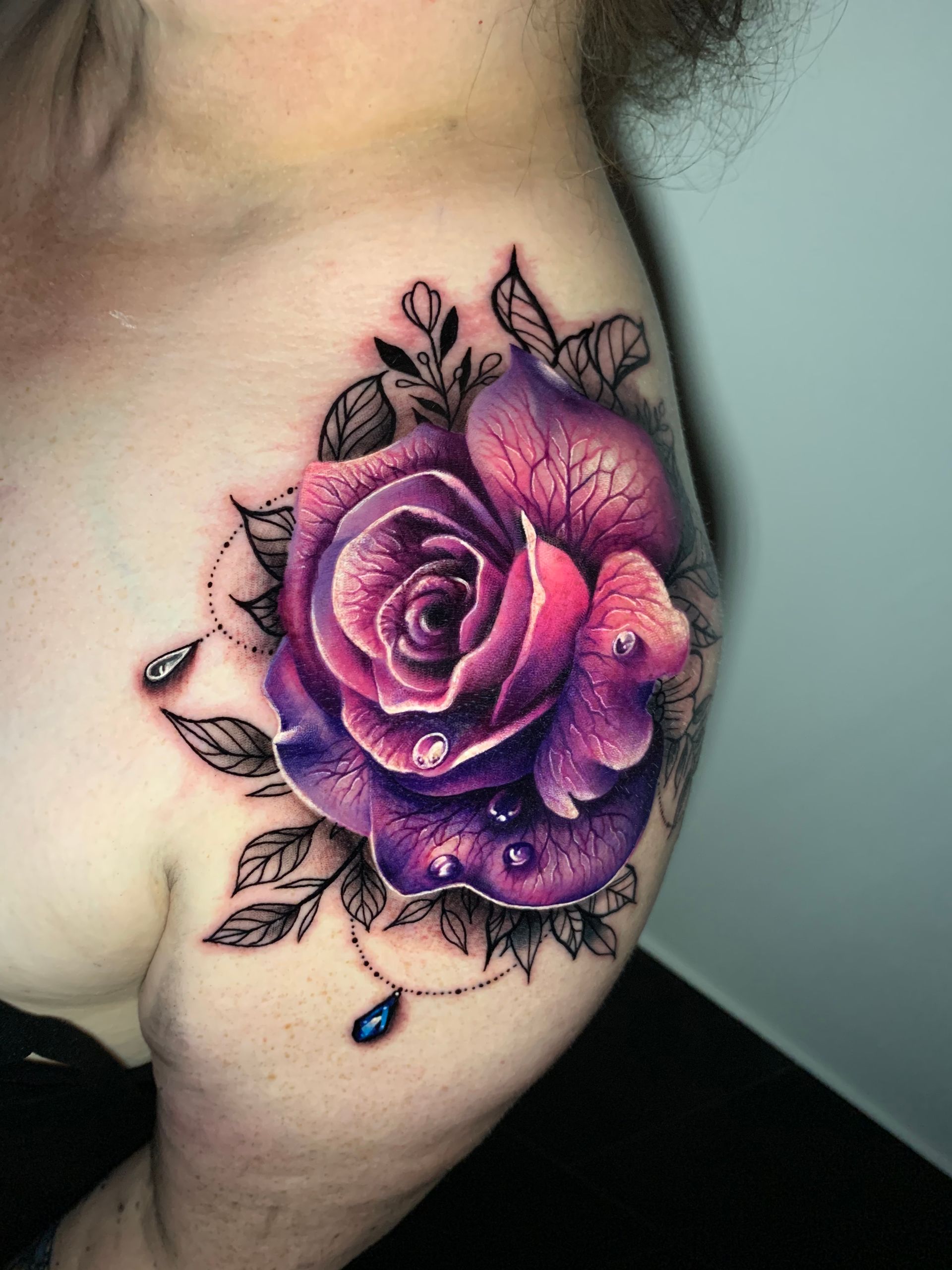 Tattoo of a vibrant purple rose on a person's shoulder, with water droplets and delicate black leaves.