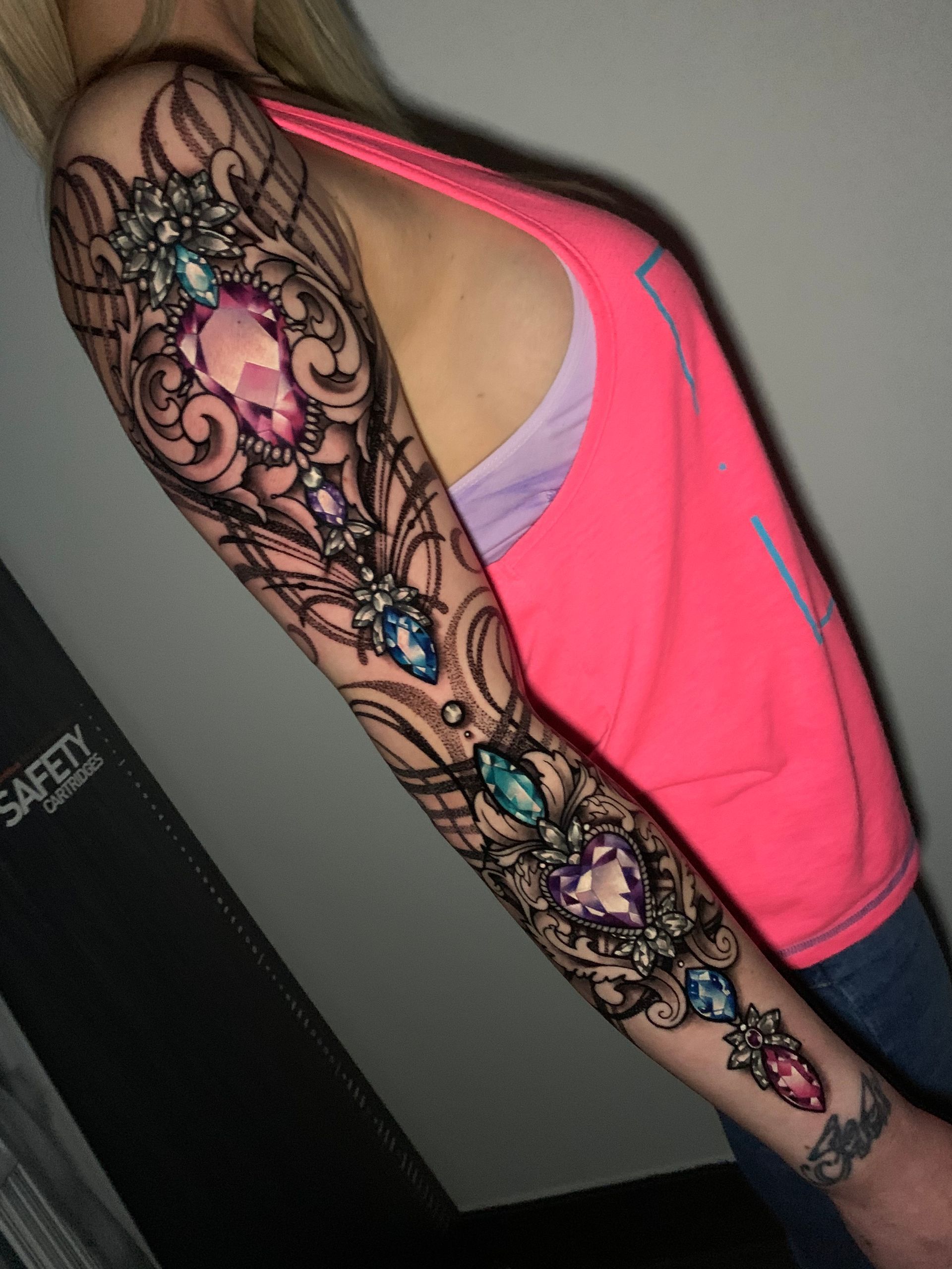 Woman's arm covered in a jeweled, ornamental tattoo featuring colorful gems and intricate black linework, with a pink tank top.