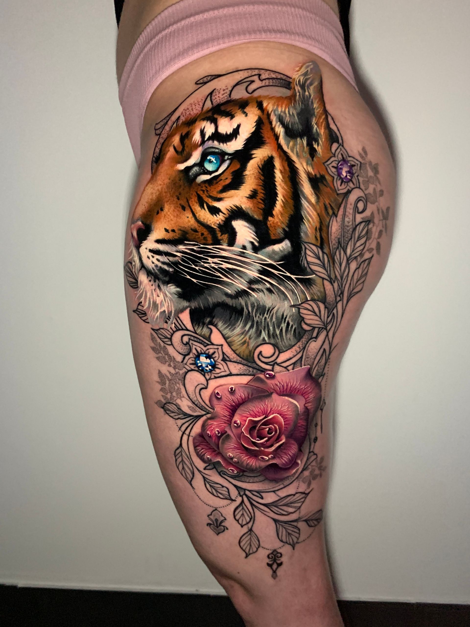 Tiger head tattoo with bright blue eyes, surrounded by a red rose and foliage on a person's thigh.