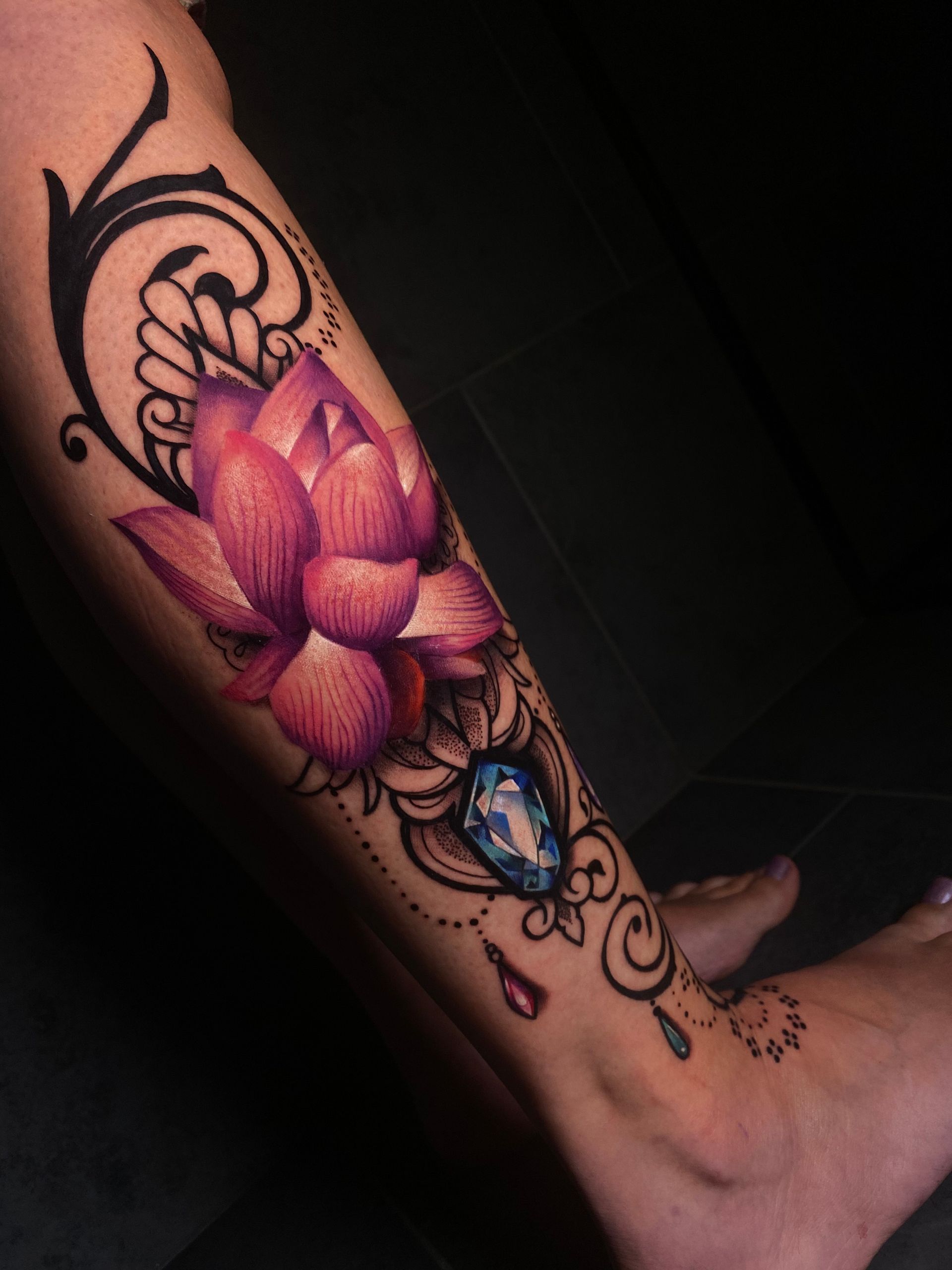 Leg tattoo featuring a pink lotus flower, blue gemstone, and ornate black flourishes.