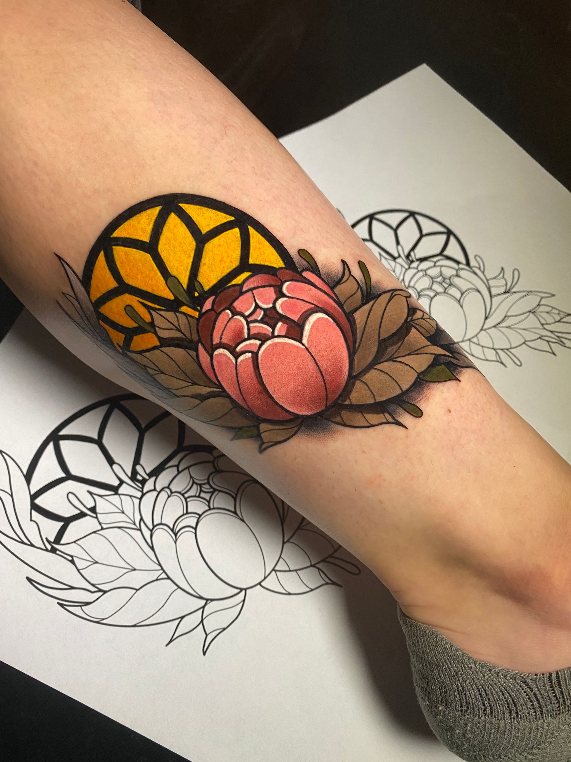 Tattoo of a pink peony with yellow stained-glass design on arm, surrounded by brown leaves. Outlines of the design are visible on paper.