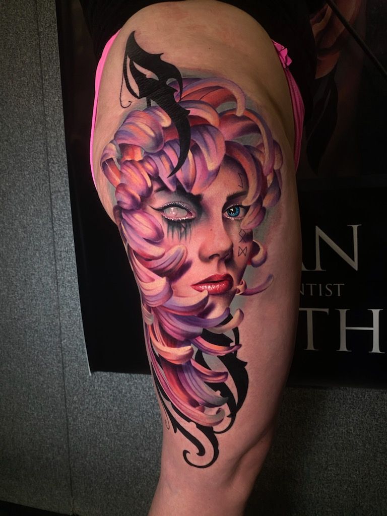 Tattoo of a woman's face with white, one blue eye surrounded by colorful flower petals on a leg.