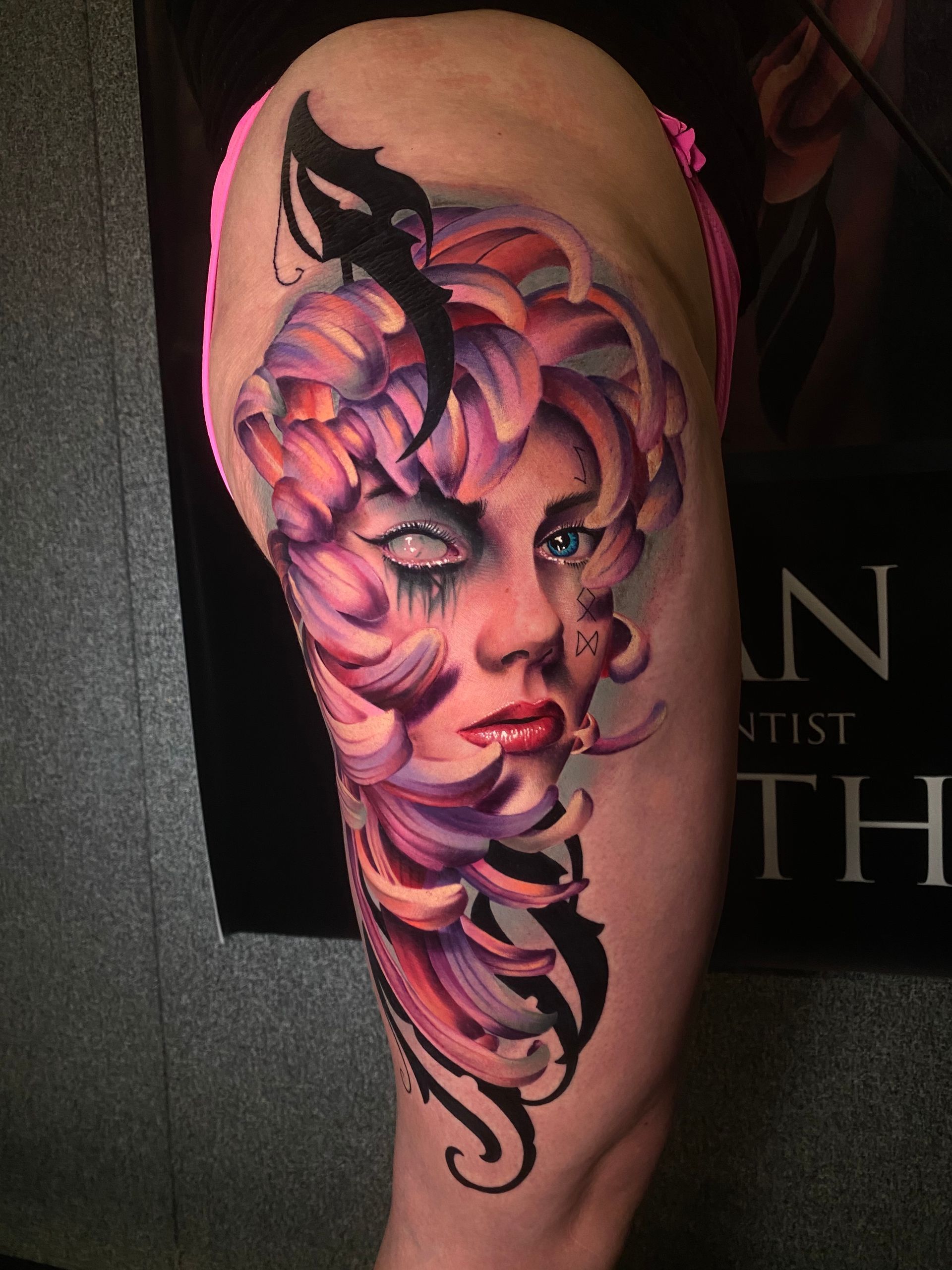Tattoo on thigh features a woman's face with pink and purple floral elements. One eye is closed with a tear, the other is open and blue.