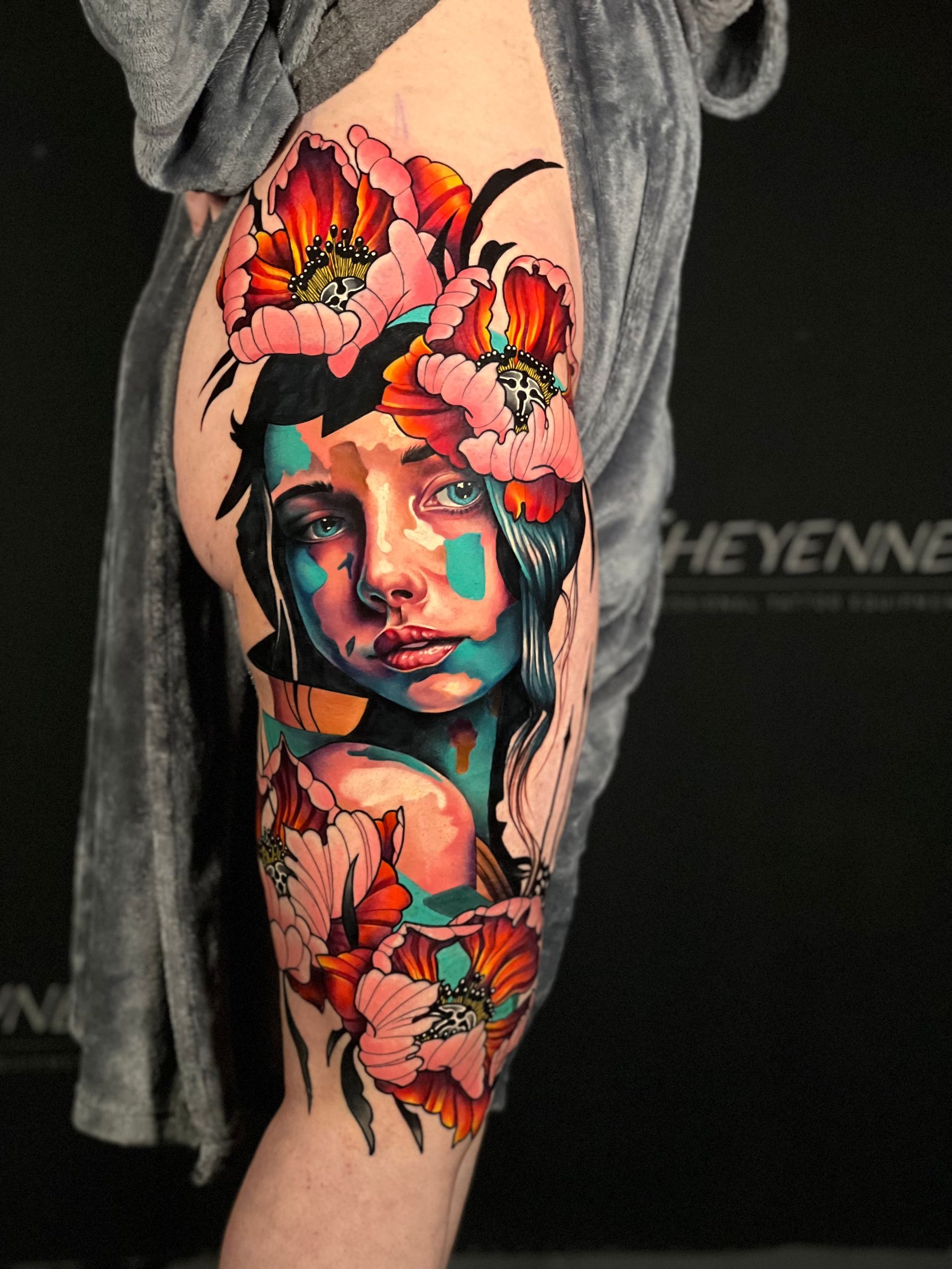 Tattoo of a woman's face and flowers in vibrant colors covering a thigh. Woman's expression is somber.
