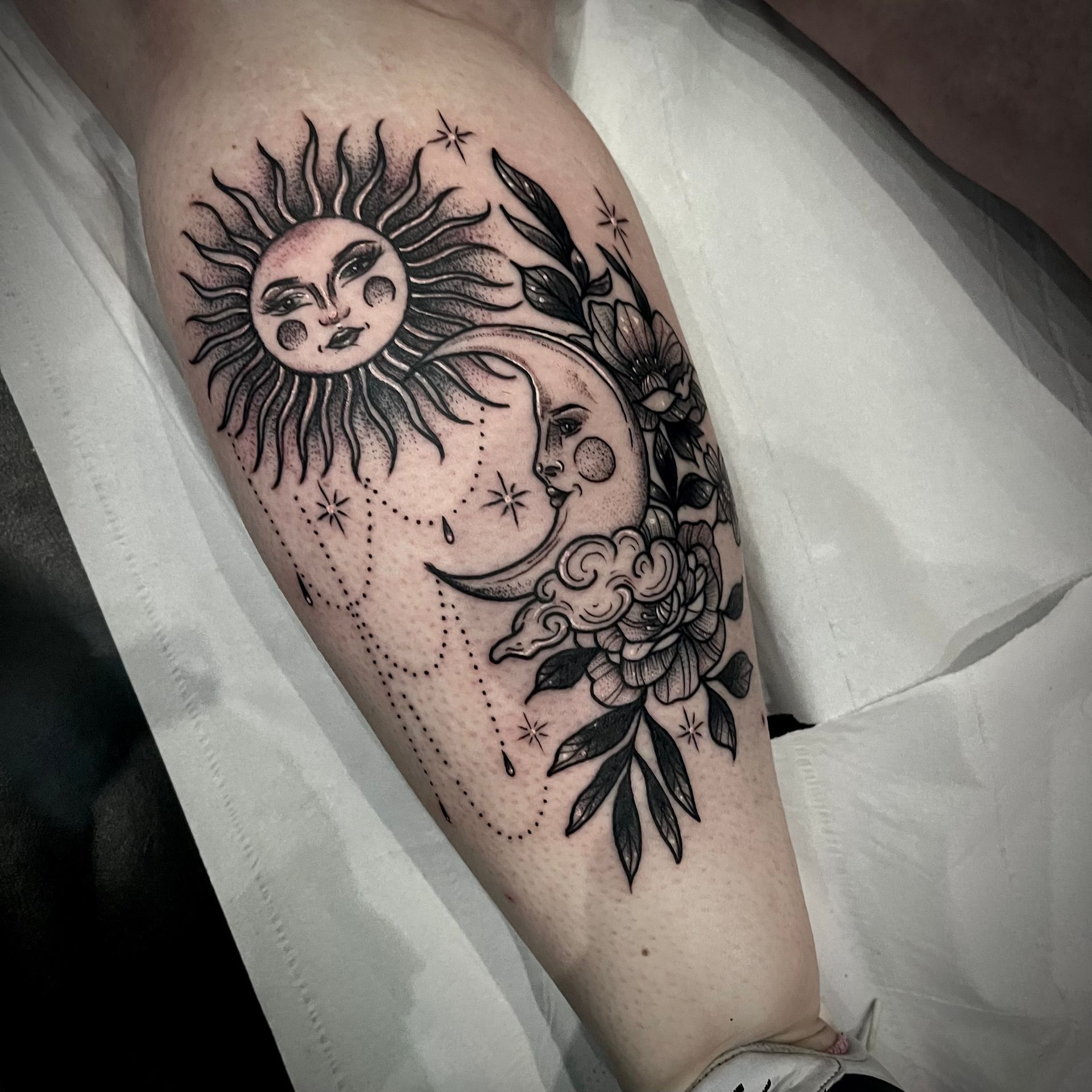 Black tattoo on an arm featuring a smiling sun, moon, stars, and flowers.